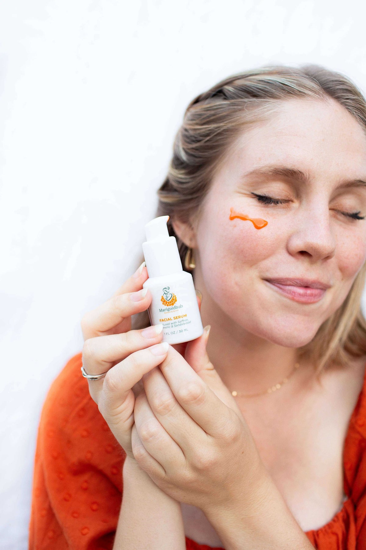 Illuminating Face Serum in a small white pump bottle with orange branding, held beside a woman's face with a serum dab