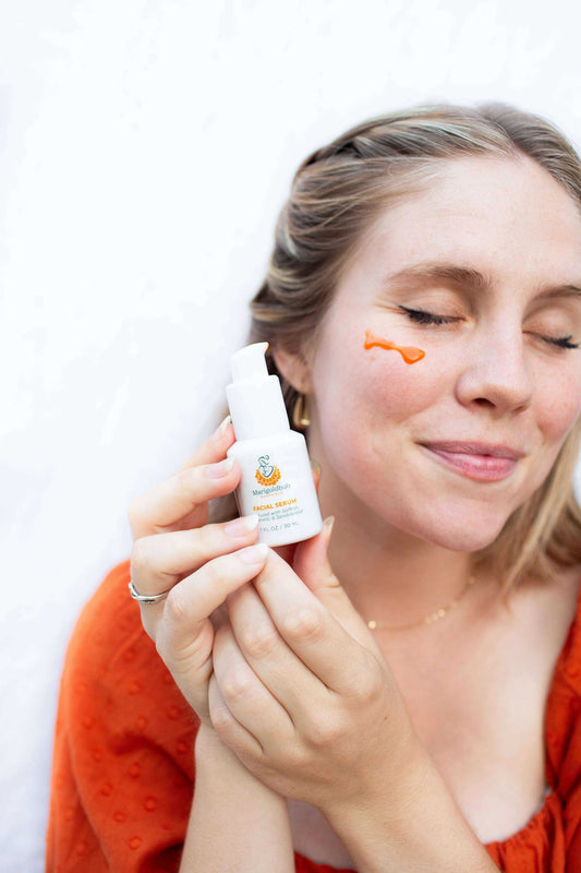 Illuminating Face Serum in a small white pump bottle with orange branding, held beside a woman's face with a serum dab