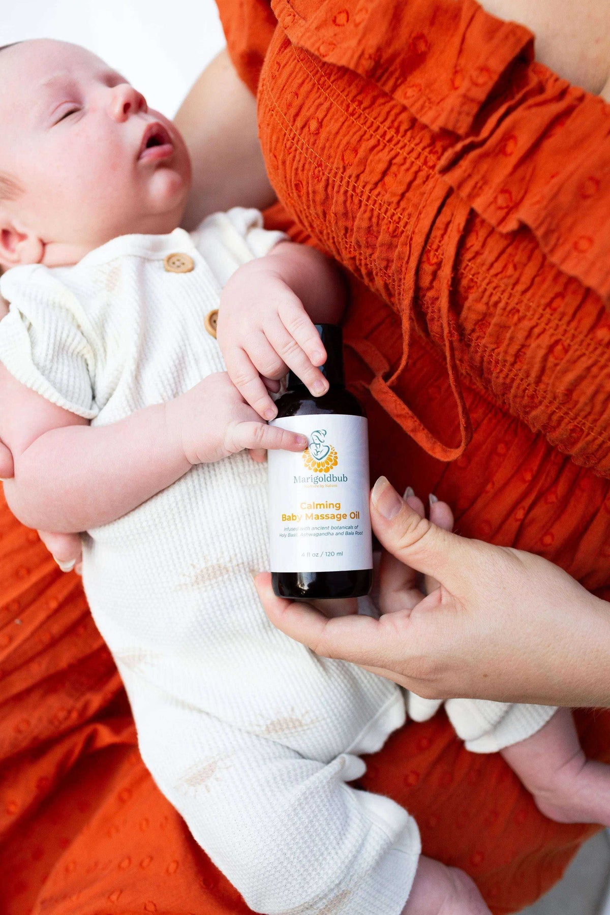 Calming Baby Massage Oil in a dark bottle with a white label, held beside a baby wearing a white outfit