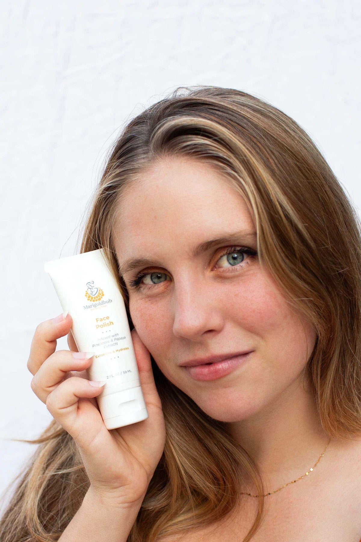 Pineapple and Papaya Skin Polish in a white tube held beside a woman’s face, with a small gold botanical logo