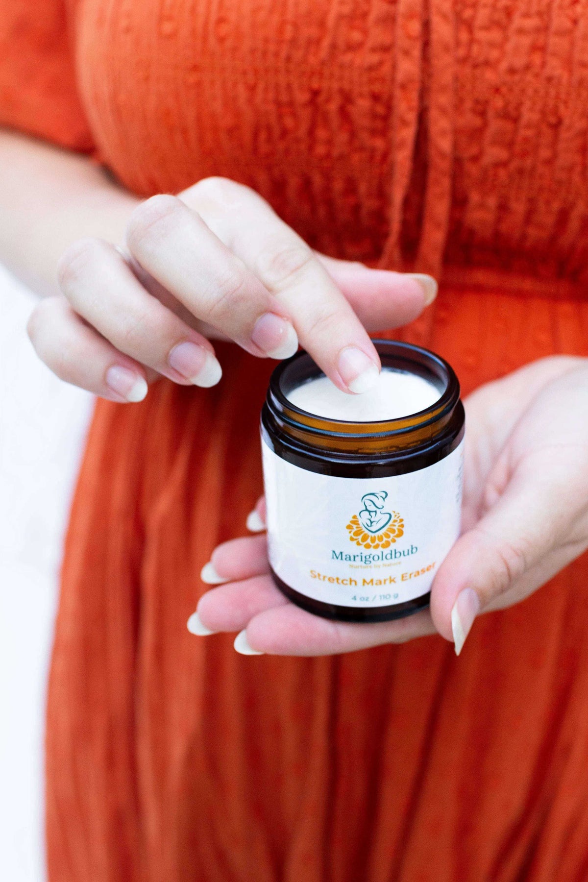 Stretch Mark Eraser cream in a brown jar with a white label, shown with a finger touching the cream